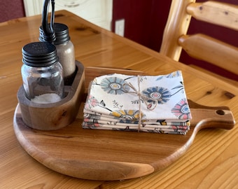 May include: A wooden serving tray with a handle holds a set of floral print napkins tied with twine, and a wooden caddy with glass jars for salt and pepper. The tray and caddy are made of natural wood.