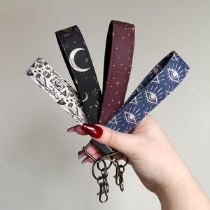 May include: Five different patterned keychains with a silver key ring. The keychains are made of fabric and feature various designs, including stars, moons, eyes, and botanical illustrations.