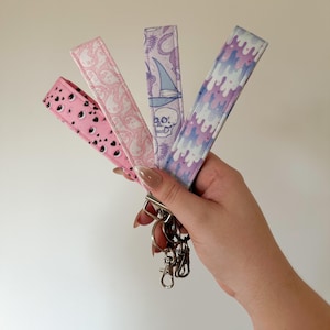 May include: Four pink and purple keychains with different patterns. The keychains feature a skull, eyes, a witch, and a dripping pattern. The keychains are attached to a silver key ring.