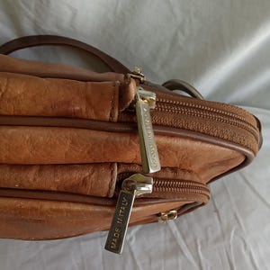 Puede incluir: Un bolso bandolera de cuero marr&oacute;n con m&uacute;ltiples compartimentos y cierre de cremallera. El bolso tiene una correa para el hombro y tiradores de cremallera met&aacute;licos. Los tiradores de la cremallera tienen la inscripci&oacute;n "MADE IN ITALY".