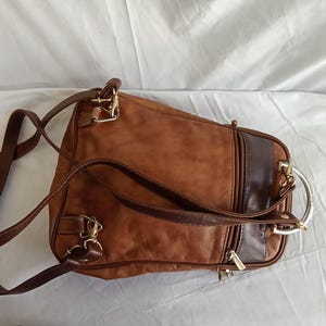 Puede incluir: Una mochila de cuero marr&oacute;n con correas ajustables y herrajes dorados. La bolsa presenta un panel de acento marr&oacute;n oscuro y un asa de anillo de metal plateado. La bolsa est&aacute; dise&ntilde;ada con m&uacute;ltiples compartimentos y cremalleras.