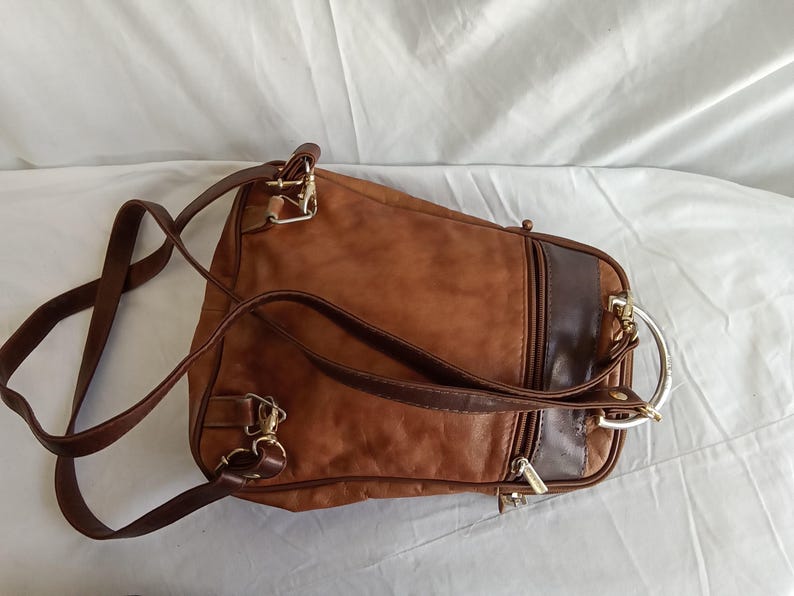 Puede incluir: Una mochila de cuero marr&oacute;n con correas ajustables y herrajes dorados. La bolsa presenta un panel de acento marr&oacute;n oscuro y un asa de anillo de metal plateado. La bolsa est&aacute; dise&ntilde;ada con m&uacute;ltiples compartimentos y cremalleras.