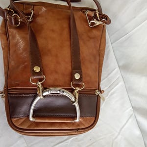 Puede incluir: Bolso de hombro de cuero marr&oacute;n con una secci&oacute;n marr&oacute;n oscuro en la parte inferior. El bolso tiene un asa plateada y el nombre "Valentino di Paolo" es visible. Tiene herrajes dorados y una cremallera.