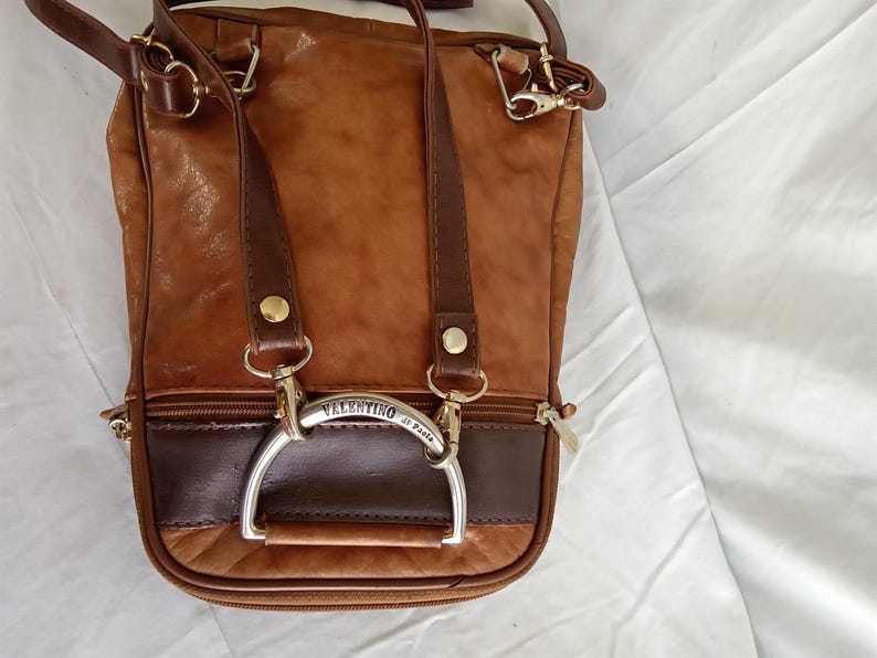 Puede incluir: Bolso de hombro de cuero marr&oacute;n con una secci&oacute;n marr&oacute;n oscuro en la parte inferior. El bolso tiene un asa plateada y el nombre "Valentino di Paolo" es visible. Tiene herrajes dorados y una cremallera.