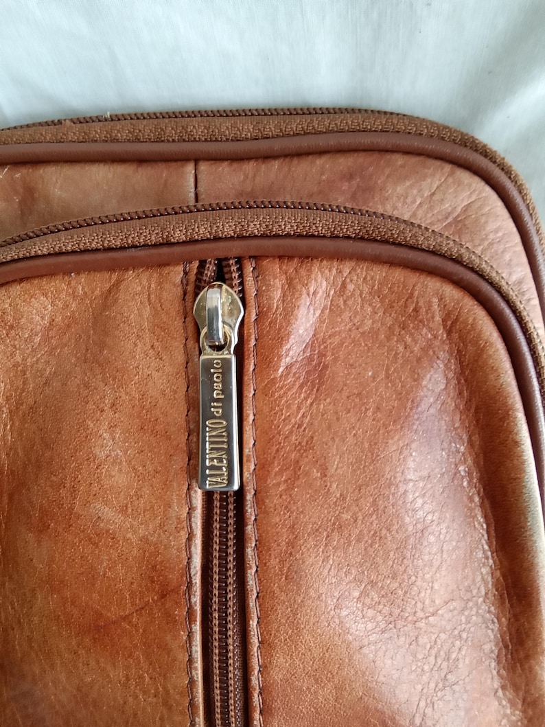 Puede incluir: Primer plano de un bolso de cuero marr&oacute;n con cremallera. El tirador de la cremallera tiene el texto "VALENTINO di paolo". El bolso tiene una superficie texturizada y una cremallera marr&oacute;n.