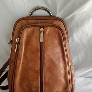 Puede incluir: Una mochila de cuero marr&oacute;n con asa superior y cremallera frontal. La mochila tiene forma redondeada y un asa met&aacute;lica con la palabra "Valentino". La bolsa tiene un bolsillo frontal con cremallera y correas marrones.