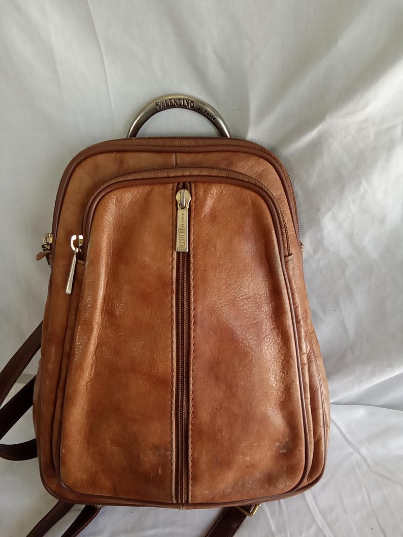 Puede incluir: Una mochila de cuero marr&oacute;n con asa superior y cremallera frontal. La mochila tiene forma redondeada y un asa met&aacute;lica con la palabra "Valentino". La bolsa tiene un bolsillo frontal con cremallera y correas marrones.