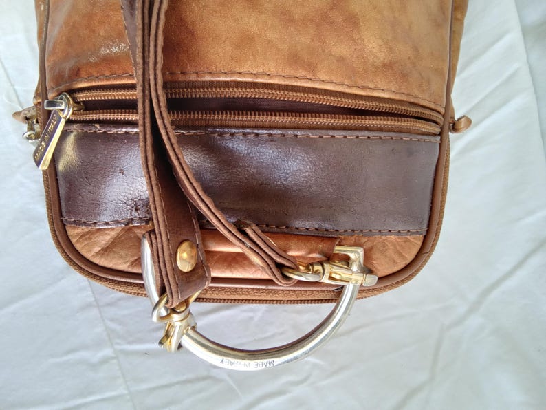 Puede incluir: Primer plano de un bolso de cuero marr&oacute;n con un panel de acento marr&oacute;n oscuro. El bolso tiene un cierre de cremallera, un asa de metal plateado y una correa. Las palabras "MADE IN ITALY" son visibles en el asa.
