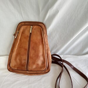 Puede incluir: Una mochila vintage de cuero marr&oacute;n con forma rectangular y cremallera vertical. La bolsa tiene una correa larga y fina de color marr&oacute;n y cremalleras doradas. El cuero muestra signos de desgaste.
