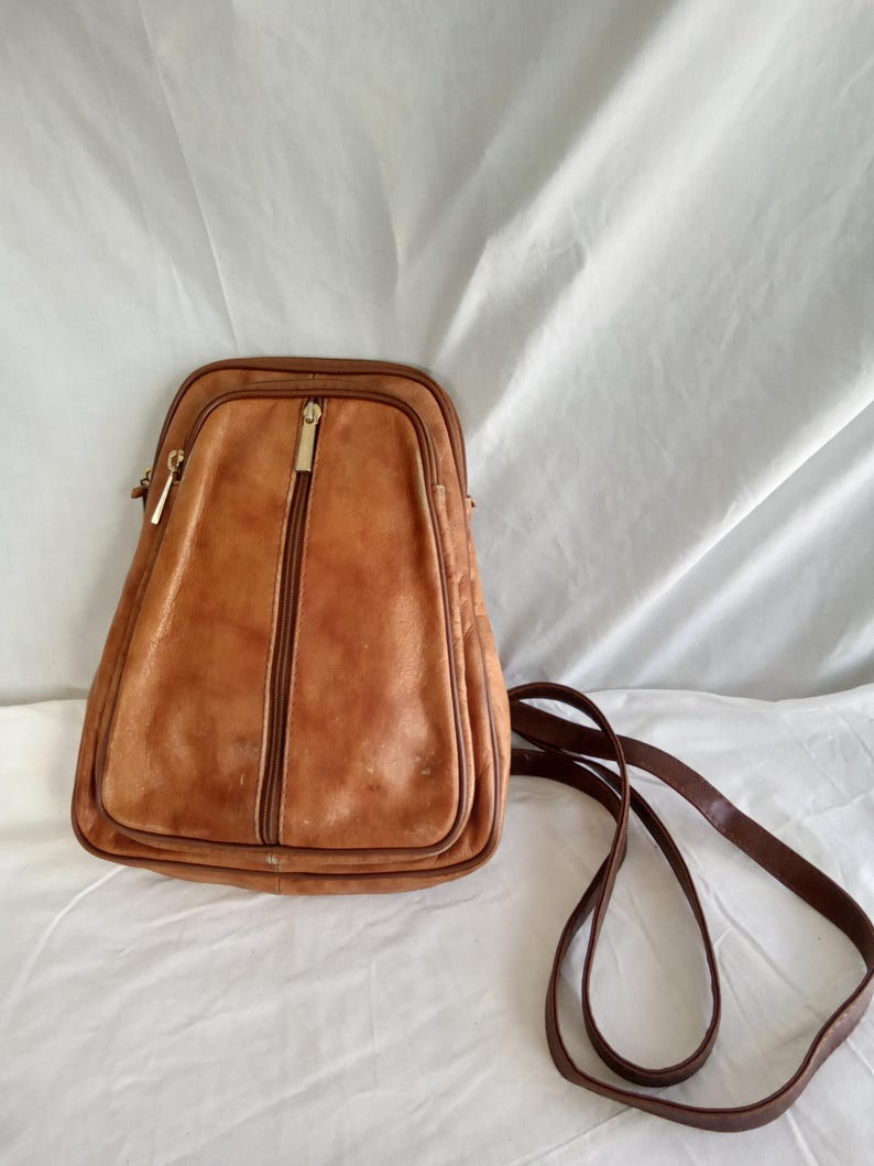 Puede incluir: Una mochila vintage de cuero marr&oacute;n con forma rectangular y cremallera vertical. La bolsa tiene una correa larga y fina de color marr&oacute;n y cremalleras doradas. El cuero muestra signos de desgaste.