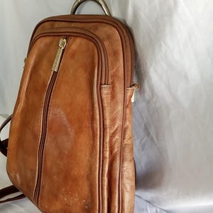 Puede incluir: Una mochila de cuero marr&oacute;n con un asa curva de metal. La mochila tiene una cremallera vertical y un bolsillo frontal m&aacute;s peque&ntilde;o. La marca "Valentino" es visible en el tirador de la cremallera. La bolsa tiene correas marrones.
