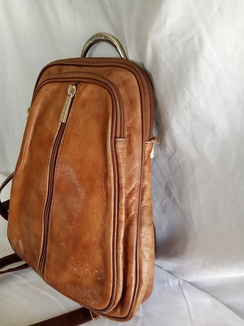 Puede incluir: Una mochila de cuero marr&oacute;n con un asa curva de metal. La mochila tiene una cremallera vertical y un bolsillo frontal m&aacute;s peque&ntilde;o. La marca "Valentino" es visible en el tirador de la cremallera. La bolsa tiene correas marrones.