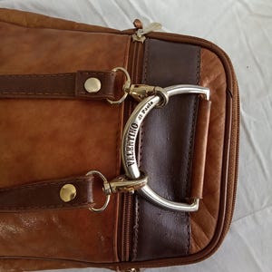 Puede incluir: Primer plano de un bolso de cuero marr&oacute;n con un asa plateada. El bolso tiene un cierre de cremallera y dos correas de cuero marr&oacute;n con remaches dorados. El asa est&aacute; grabada con las palabras "Valentino di Paolo".