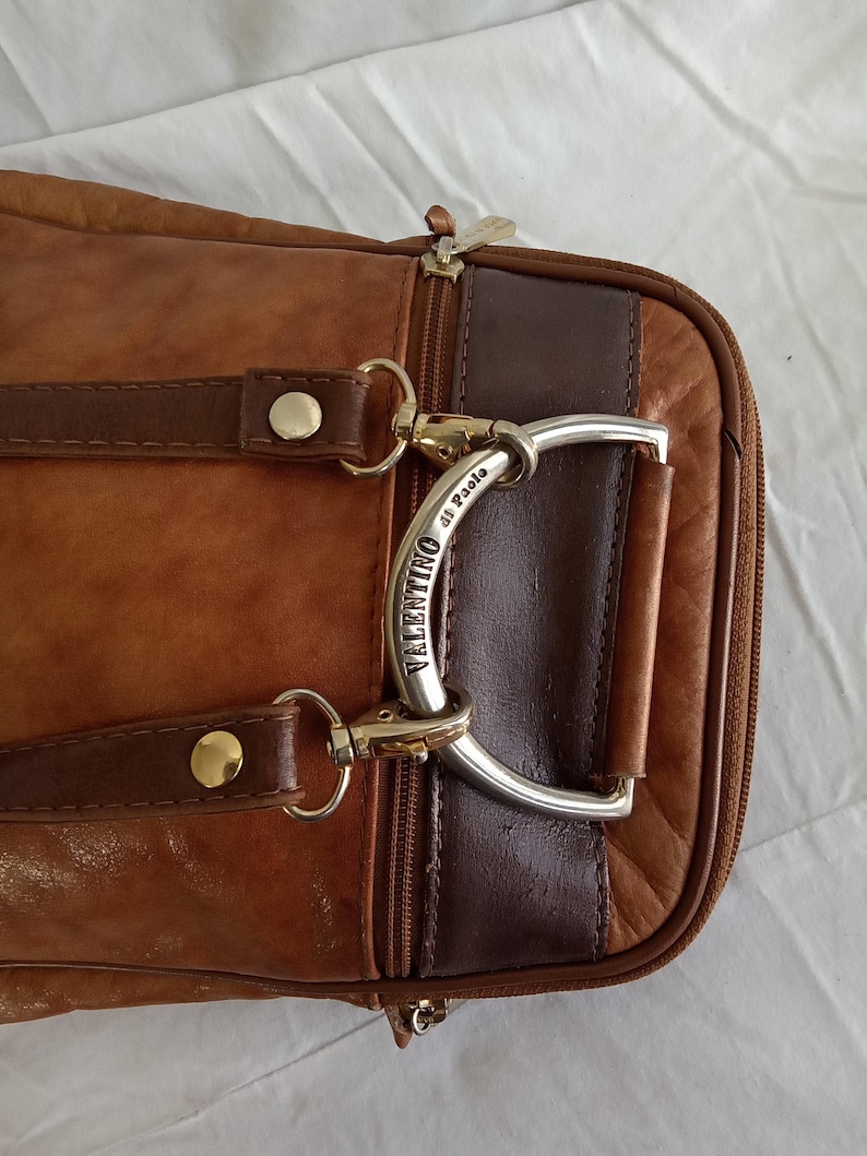 Puede incluir: Primer plano de un bolso de cuero marr&oacute;n con un asa plateada. El bolso tiene un cierre de cremallera y dos correas de cuero marr&oacute;n con remaches dorados. El asa est&aacute; grabada con las palabras "Valentino di Paolo".