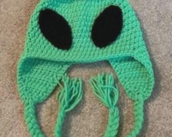 May include: A handmade, green crocheted alien hat with large black oval eyes. The hat features ear flaps and braided ties with fringe. This whimsical hat is perfect for costume parties or everyday wear.