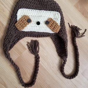 May include: A handmade crochet hat in the shape of a sloth. The hat is primarily brown with a white face, brown accents, and black button eyes. Braided ties with tassels hang from the ear flaps. A whimsical and cozy accessory.