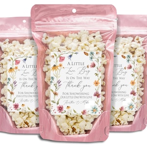 May include: Three pink cellophane bags filled with popcorn. Each bag has a floral label with the text "A Little Love Bug Is On The Way Thank You". The bags are sealed at the top and filled with white popcorn.