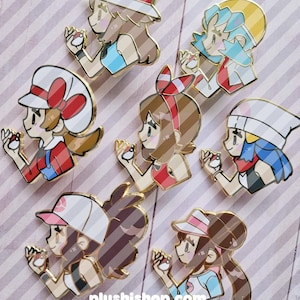 May include: A collection of seven enamel pins featuring stylized anime-style characters, each wearing a unique hat and holding a small Poké Ball. The pins have gold-toned metal outlines and vibrant colors, including red, blue, white, and brown accents.