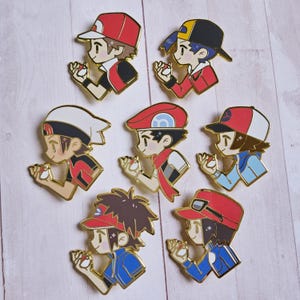 May include: A collection of eight enamel pins featuring various cartoon characters in dynamic poses. Each pin has a gold-colored outline and depicts a character holding a small, round object. The characters wear hats and outfits in red, blue, and white.