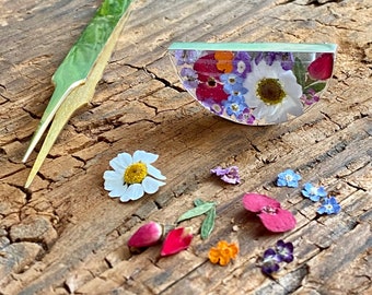 May include: A half-circle resin piece featuring a selection of pressed flowers in shades of pink, purple, white, and yellow. The resin piece is resting on a wooden surface with loose flowers scattered around it. A pair of gold tweezers is also visible in the image.