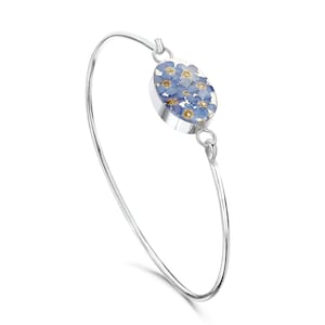 May include: A silver bangle bracelet with a round charm featuring pressed forget-me-not flowers in blue and yellow.