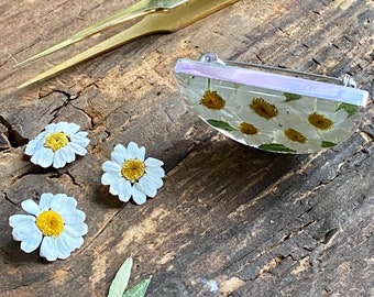 May include: A silver and white resin brooch in a half-circle shape. The brooch is filled with pressed white daisies. Three loose white daisies are on a wooden surface next to the brooch.