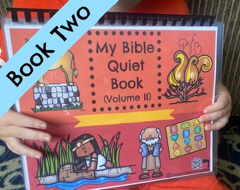 May include: A child holds a red and black spiral bound book titled "My Bible Quiet Book (Volume II)". The book has illustrations of a camel, a burning bush, a woman in a boat, and a man with a beard.