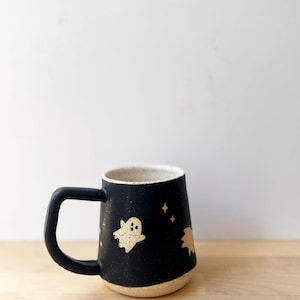 May include: Black ceramic mug with a cream-colored interior and base. The mug features a black handle and is decorated with beige ghost and star designs. The mug is approximately 4 inches tall.