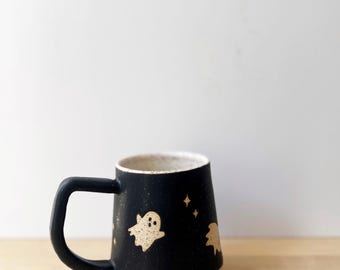 May include: Black ceramic mug with a cream-colored interior and base. The mug features a black handle and is decorated with beige ghost and star designs. The mug is approximately 4 inches tall.