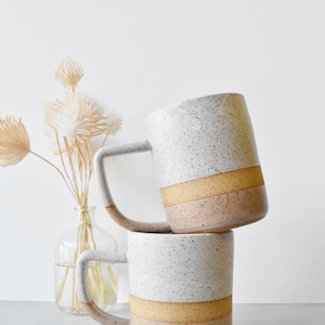 May include: Two ceramic mugs stacked on top of each other. The mugs have a speckled white upper section, a yellow-tan middle band, and a light pink lower band. The handles are a light gray color. A vase of dried flowers is in the background.