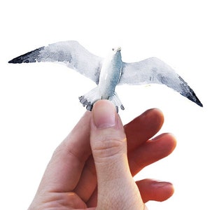 Peut inclure: Une peinture à l'aquarelle d'une mouette avec ses ailes déployées, tenue dans la main d'une personne.