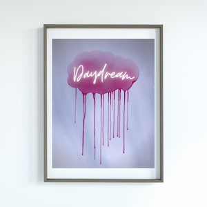 May include: Framed art print with a pink cloud and dripping paint effect on a light blue background. The word "Daydream" is written in a neon-style font across the cloud. The frame is a neutral grey colour.