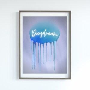 May include: Framed art print with the word "Daydream" in white script over a blue watercolor cloud with dripping paint effect. The artwork is set against a soft, light purple background and framed in a grey wooden frame.