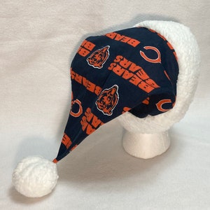 Op de afbeelding: Een witte en blauwe kerstmanmuts met een witte pompon. De muts is gemaakt van fleece en heeft een Chicago Bears logo op de voorkant.