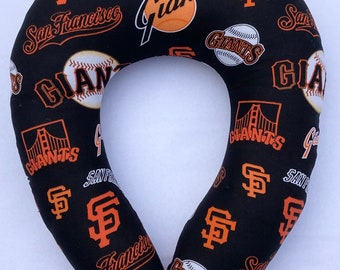 May include: Black travel pillow with an orange and white San Francisco Giants logo pattern. The pillow is shaped like a horseshoe and has a hole in the middle for the neck.