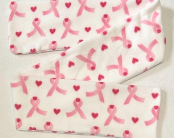 May include: A white fleece headband featuring a repeating pattern of pink awareness ribbons and small red hearts. The headband is folded and lies flat, showcasing the design. The soft material suggests comfort and warmth.