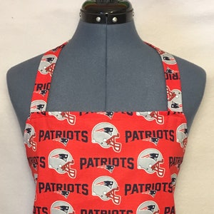 以下が含まれることがあります： 白のフットボールヘルメットと黒の「Patriots」という文字が繰り返しパターンで描かれた赤いキッチンエプロンです。