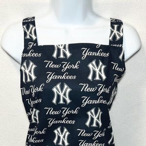以下が含まれることがあります： 白のニューヨーク・ヤンキースのロゴと白の筆記体で書かれた「New York Yankees」の文字が全体にプリントされた、ダークネイビーブルーのエプロン。スクエアネックで、幅の広いショルダーストラップが2本付いています。