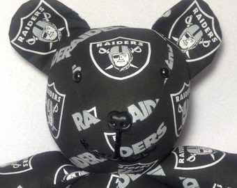 May include: Black and silver teddy bear with the Oakland Raiders logo repeated on the fabric. The bear has black button eyes and a black nose.