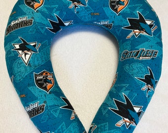 May include: A teal blue travel neck pillow with a repeating pattern of the San Jose Sharks NHL team logo. The logo features a shark with a hockey stick and the words "San Jose Sharks".