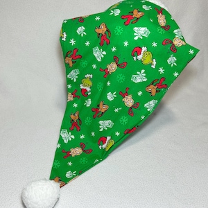 Könnte beinhalten: Ein grüner Weihnachtsmannhut mit einem weißen Bommel und weißem Kunstfellbesatz. Der Hut ist mit Zeichentrickfiguren, Schneeflocken und verpackten Geschenken gemustert. Die Figuren sind in Rot, Braun und Grün gehalten.
