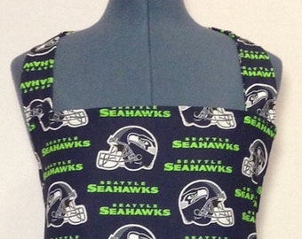 May include: A blue and green apron with a repeating pattern of Seattle Seahawks logos and helmets. The straps are also patterned with the team logo.