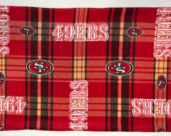 May include: Red plaid blanket featuring the San Francisco 49ers logo and team name in white. The blanket has a repeating pattern of the team's logo and name, with a red, yellow, and green plaid design.