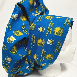Peut inclure: Un bandeau en tissu bleu et or avec le logo des Golden State Warriors répété sur toute la surface. Le tissu est plié et noué pour créer un couvre-chef de style turban.