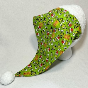 May include: A green and white Santa hat with a Grinch-themed pattern. The hat has a white fluffy trim and a white pom-pom at the tip. The Grinch design includes the text "Feelin' Grinchy".