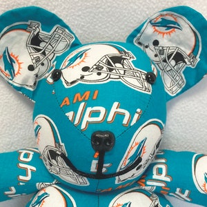 May include: A blue and orange teddy bear with a Miami Dolphins logo on its ears and chest. The bear has black button eyes and a black nose.