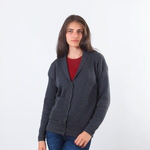 May include: A dark gray cardigan with a collared neckline and a button closure. The cardigan has a patch pocket on the left side and is made of a soft, textured fabric.