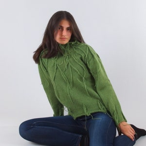 Op de afbeelding: Een groene gebreide trui met een hoge hals en lange mouwen. De trui heeft decoratieve franjes en een textuur gebreid patroon. De persoon draagt een blauwe spijkerbroek. De trui is een uniek kledingstuk.