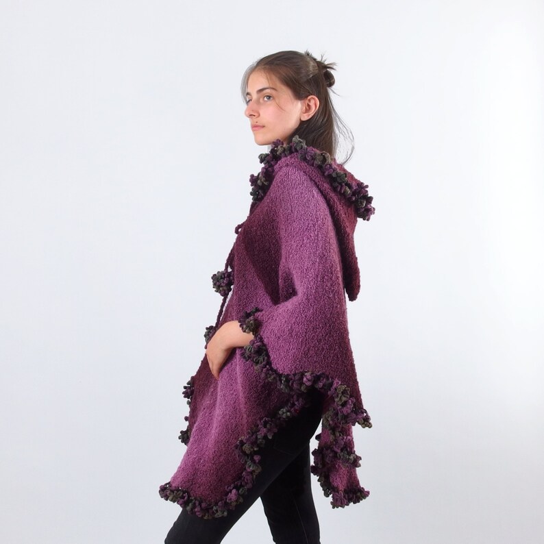 以下が含まれることがあります： フリルトリムが付いた紫色のフード付きニットポンチョ。ポンチョは柔らかくふわふわの糸でできており、ゆったりとしたフィット感です。