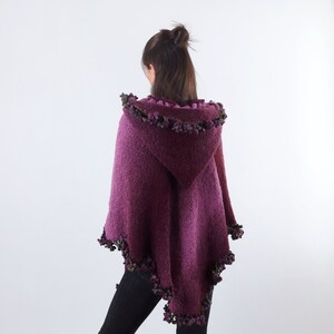 以下が含まれることがあります： 紫色のフード付きポンチョで、質感のあるニットとフリンジのトリムが付いています。フリンジは、紫と緑の色合いの小さなポンポンでできています。